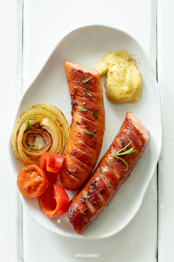 Grilled Kiełbasa (Polish Sausage) with grilled onion, cherry tomato and bell pepper, served with mustard. On a white plate, on a white wooden table. Shot from above.