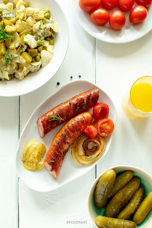 Grill Party table: Polish Grilled Kiełbasa Sausage on a white plate served with fried onions, cherry tomatoes and mustard. Dill pickles and Potato Salad. On a white wooden table, shot from above. 