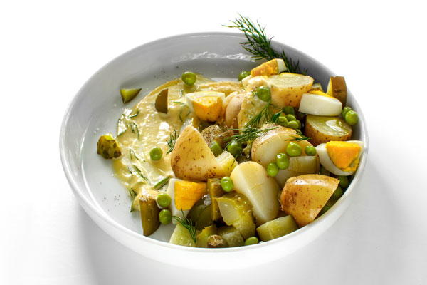 A white plate with Polish Potato Salad, made with new potatoes, dill pickles, apple, onion, green peas and mayo. On a white background, a side view.