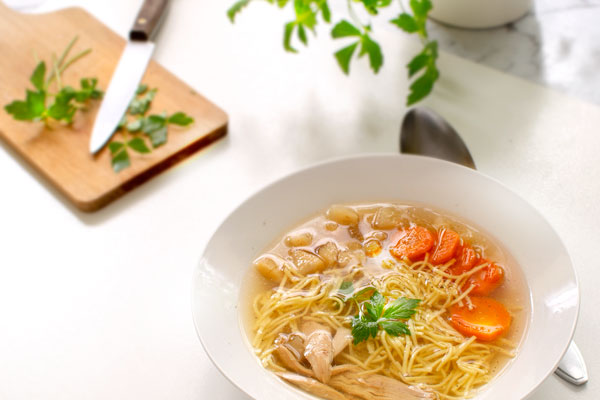 Rosół: Polish Chicken Soup with thin egg Noodles. Served with sliced carrots, parsnip root and chicken meat. Served on a white plate, on a white table. Cutting board and lovage in the distance.