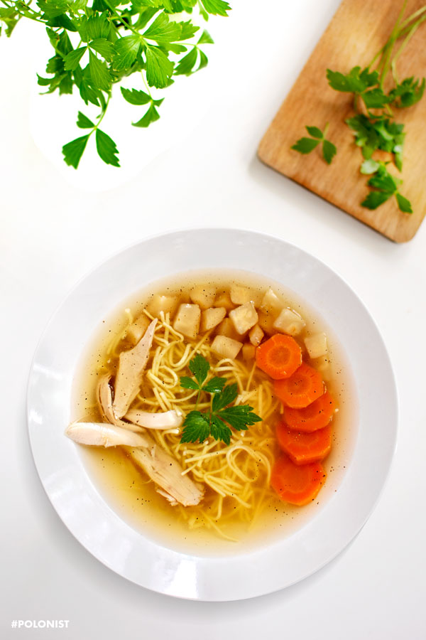 Top view of Rosół: Polish Chicken Soup with thin egg Noodles. Served with sliced carrots, parsnip root and chicken meat. Served on a white plate, on a white table. Cutting board and lovage leaves in the frame.