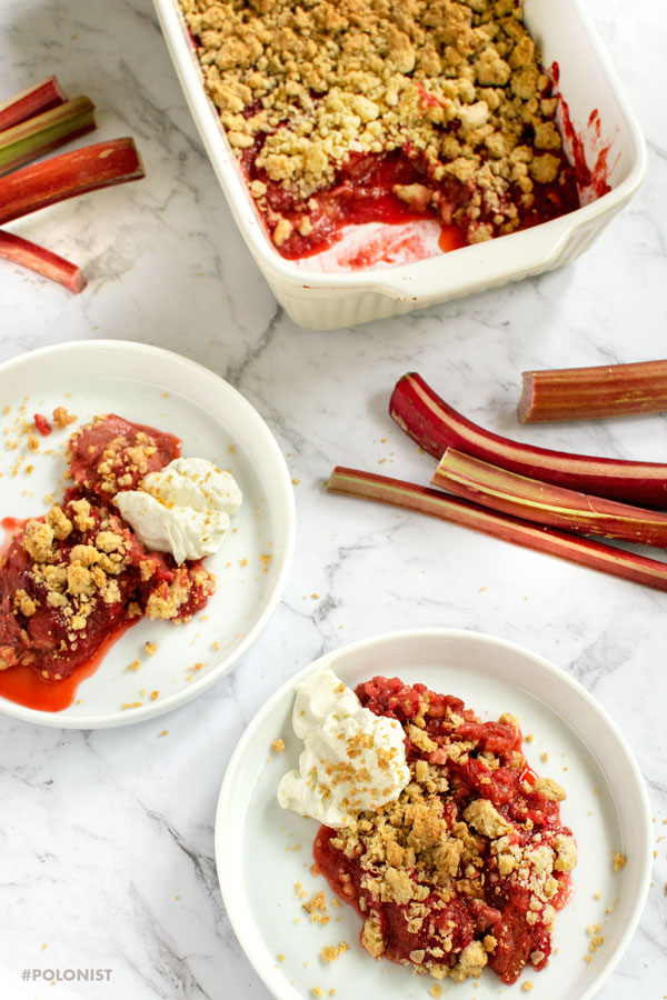 Top view/flat lay of a baking dish containing Rhubarb and Strawberry Crumble on a white marble table, together with two individual plates / potrions ,served with creamed cheese
