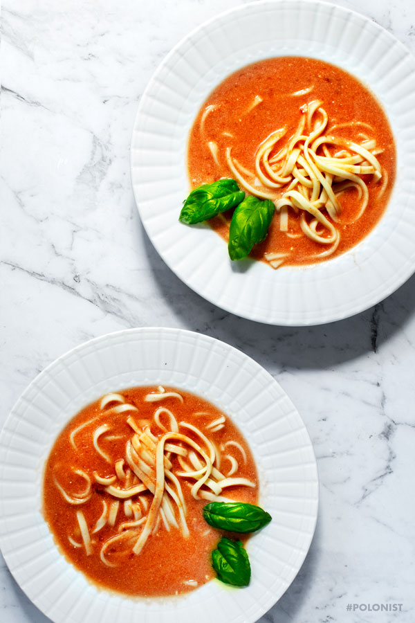 Flat lay of two white plates filled with Polish tomato Soup Pomidorowa, served with pasta, on a white marble background.