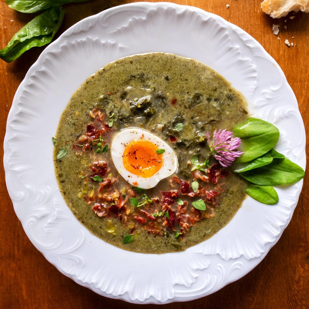 Szczawiowa: Polish Sorrel Soup, garnished with fried bacon, sorrel leaves and halved soft egg, on a wooden table.
