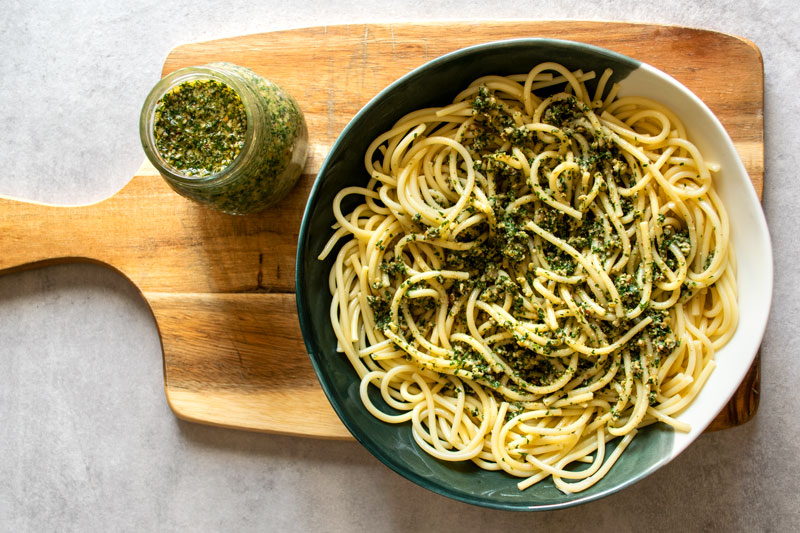 Carrot top pesto pasta, served in a bowl on top of a wooden board, with a jar of freshly made carrot leaves/greens pesto sauce.